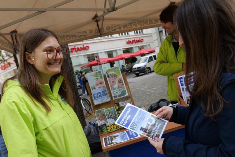 Magdalena Pitzer von GLOBAL 2000 in neon-grüner Jacke lächelt an einem Infostand, während eine Person Flyer hält