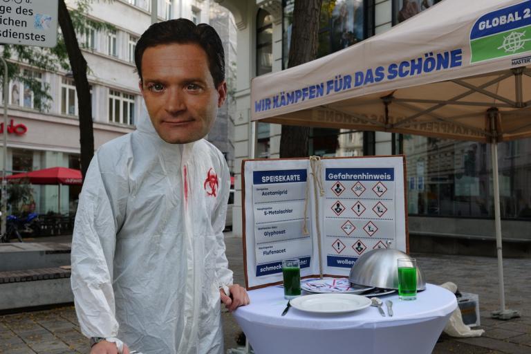 Person mit Norbert Totschnig Maske trägt weißen Schutzanzug mit Biohazard-Symbol und steht an einem Stehtisch mit gedecktem Teller und grüner Flüssigkeit vor einem Informationsstand mit Hinweisschildern zu Pestiziden