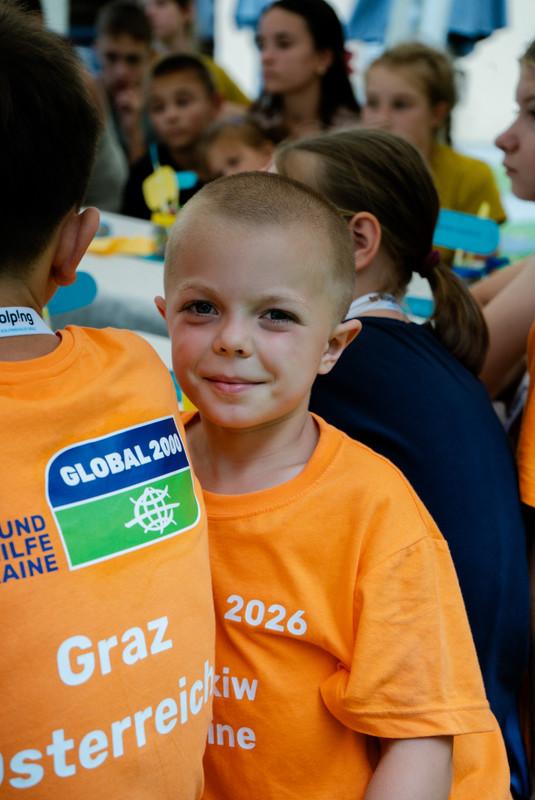 Junge mit kurzem Haar und orangefarbenem T-Shirt mit Aufdruck sitzt in einer Gruppe von Kindern