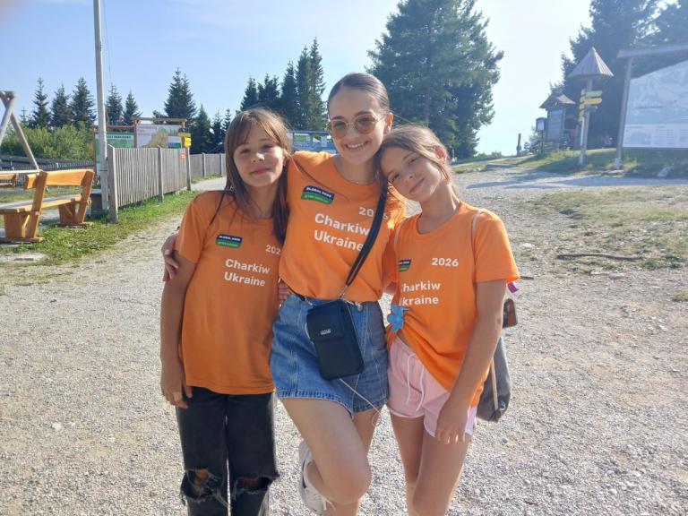Drei Mädchen in orangefarbenen T-Shirts mit der Aufschrift 'Charkiw Ukraine 2026' stehen eng umarmt auf einem sonnigen Weg mit Bäumen und Holzbänken im Hintergrund, die mittlere trägt eine Sonnenbrille und eine Umhängetasche.