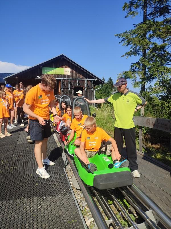 Mehrere Personen in orangefarbenen T-Shirts sitzen in einer grünen Sommerrodelbahn, umgeben von weiteren Menschen auf einem Holzsteg vor einem Holzhäuschen unter blauem Himmel