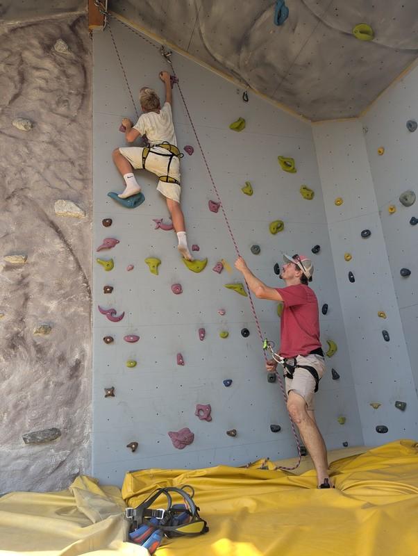 Kind klettert an einer Kletterwand mit bunten Griffen, Erwachsener sichert von unten