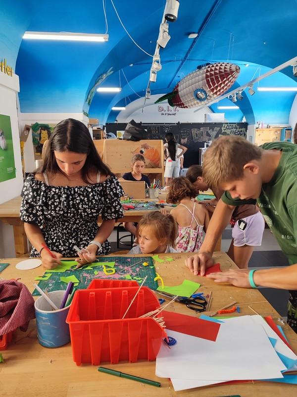 Mehrere Kinder und Jugendliche sitzen an einem Tisch und basteln mit Papier und Stiften in einem Raum mit blauem Gewölbedeckenhimmel und einer hängenden Rakete aus Pappe