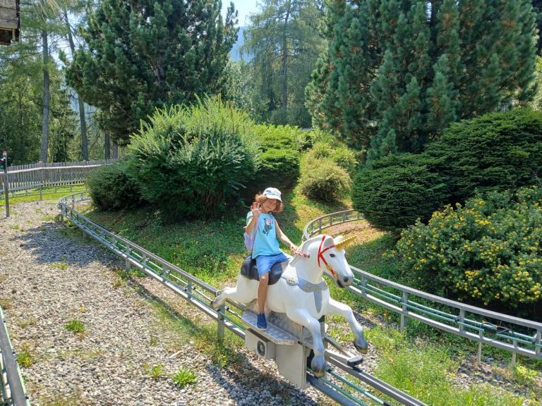 Kind mit weißem Hut sitzt auf einem weißen Einhorn-Fahrgeschäft auf einer Schiene im Freien umgeben von Büschen und Bäumen