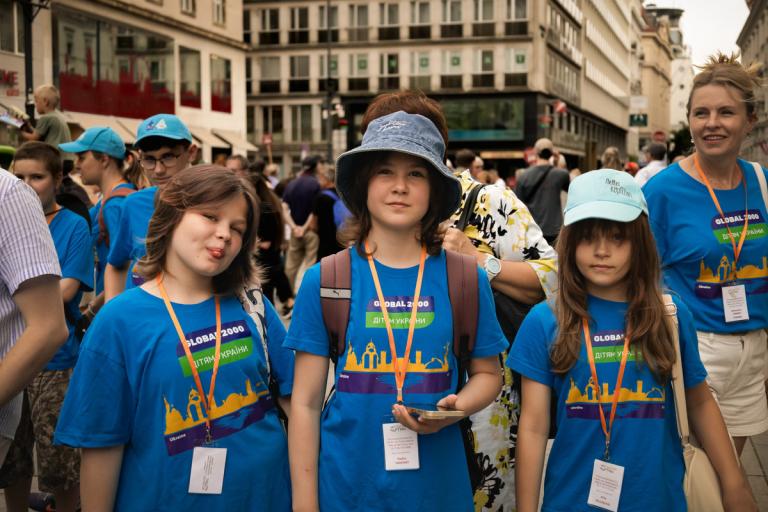 Drei Kinder mit blauen T-Shirts und Namensschildern stehen in einer Menschenmenge in einer Stadt