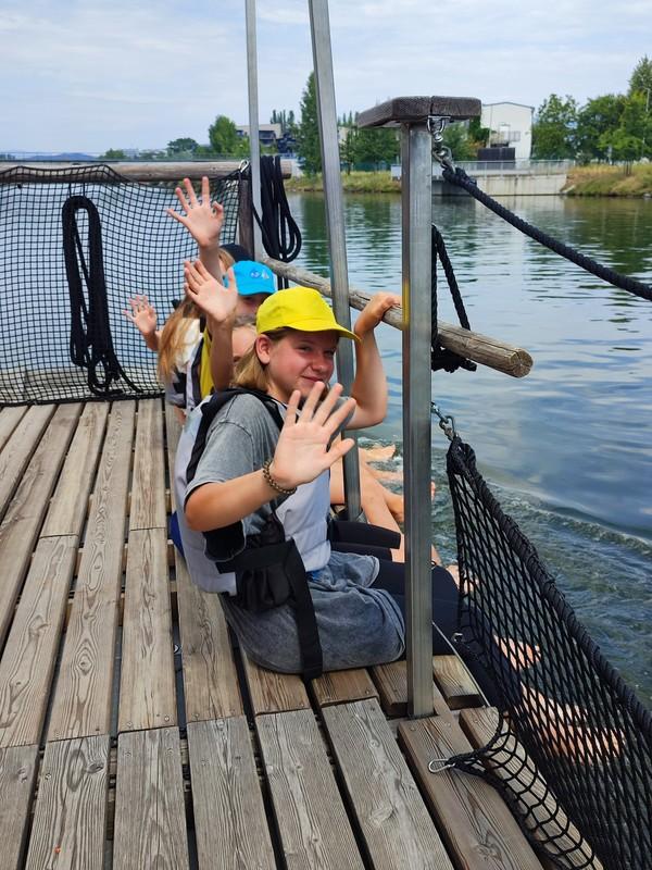Drei Jugendliche sitzen auf einem Holzsteg am Wasser und winken zur Kamera, zwei tragen bunte Kappen, einer eine gelbe