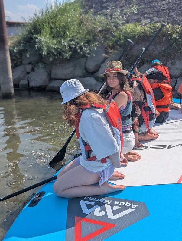 Mehrere Kinder mit Schwimmwesten knien auf einem Stand-up-Paddle-Board am Ufer