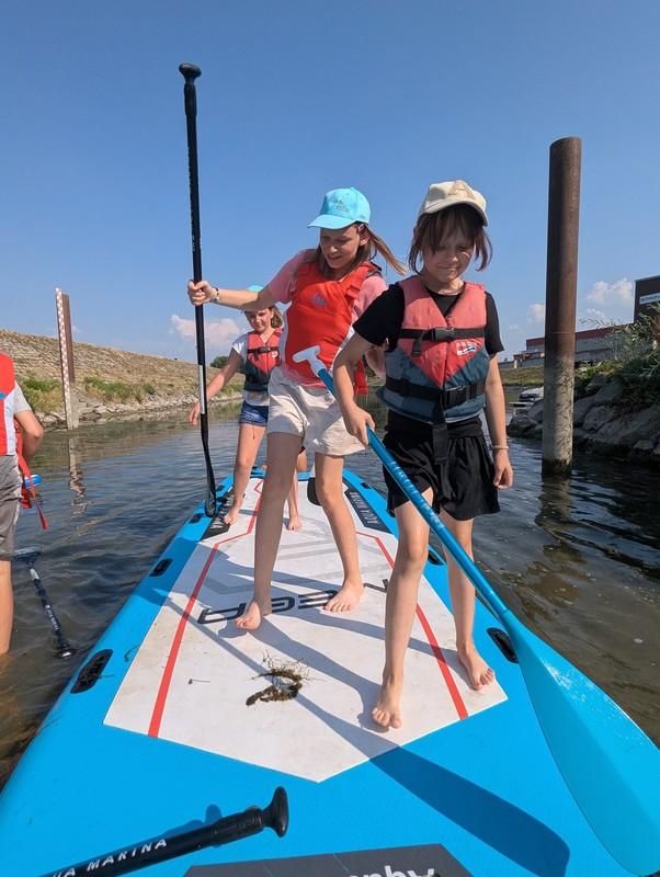 Mehrere Kinder mit Schwimmwesten stehen barfuß auf einem großen blauen Stand-up-Paddle-Board in einem schmalen Wasserkanal