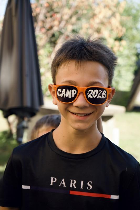 Junge mit kurzen dunklen Haaren trägt orangefarbene Sonnenbrille mit der Aufschrift 'CAMP 2026' 