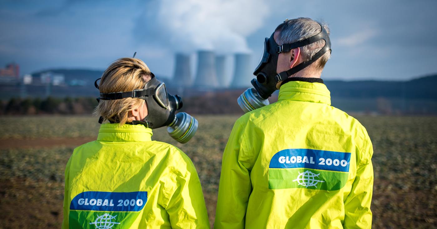 Stopp AKW Mochovce - Zwei Personen stehen vor einem AKW und sehen sich an, sie tragen GLOBAL 2000 Jacken und Schutzmasken