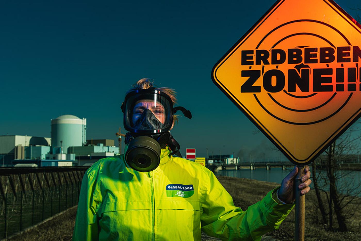 Frau mit einer Schutzmaske und einer GLOBAL 2000 Jacke steht vor dem AKW und hält ein Schild mit der Aufschrift "Erdbeben Zone!!!"