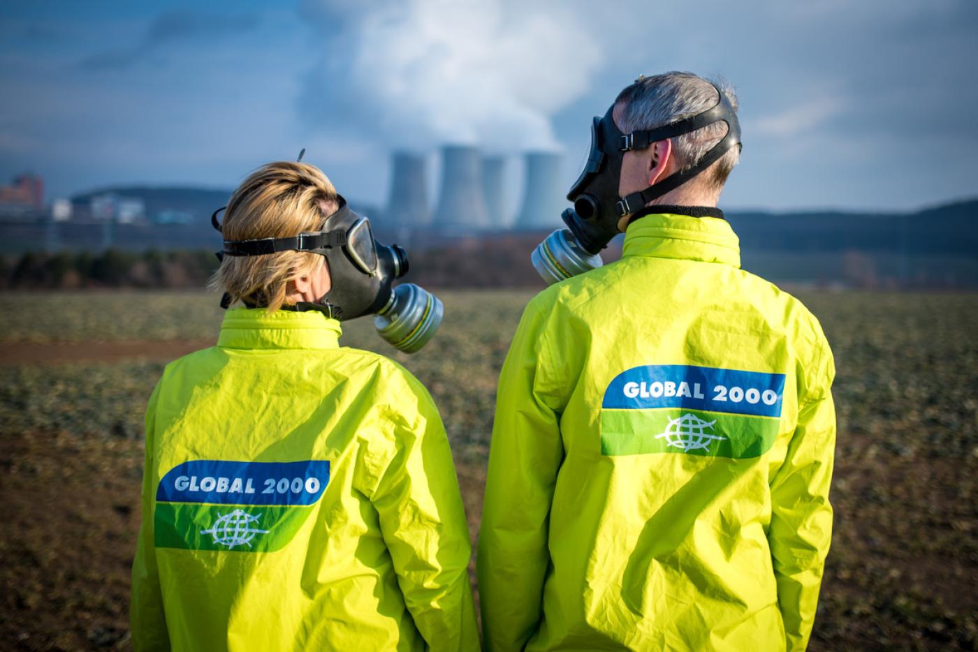 Zwei Personen stehen vor einem AKW und sehen sich an, sie tragen GLOBAL 2000 Jacken und Schutzmasken