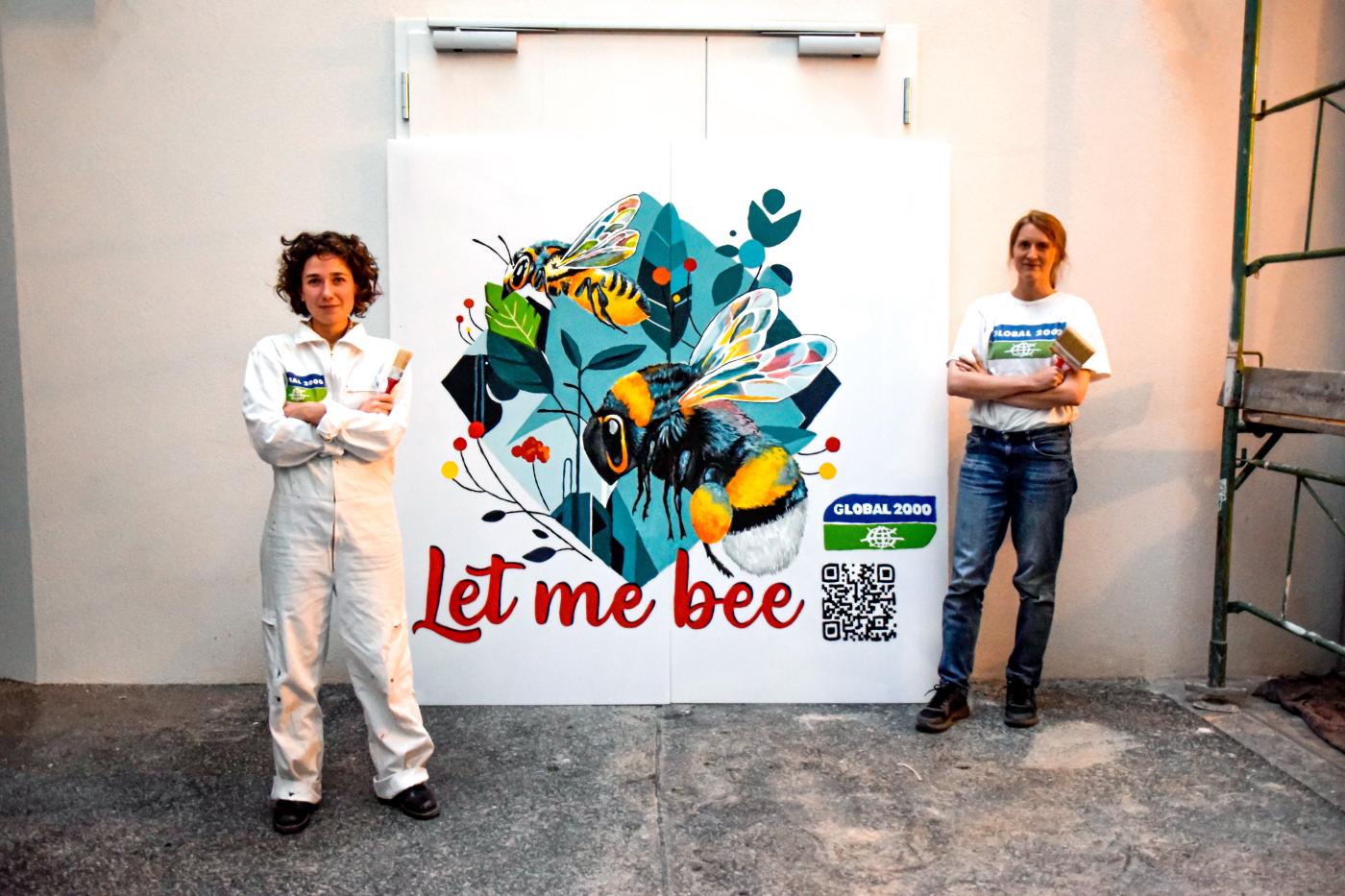 zwei Aktivistinnen mit Pinsel in den Händen stehen vor dem Bild, dass sie gerade gemalt haben. Darauf zu sehen sind zwei Bienen und der Schriftzug "Let me bee"