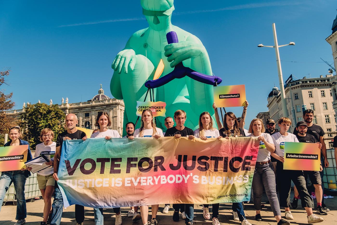 Aktivist:innen halten Banner in der Hand, worauf steht: "Vote for Justice". Sie stehen vor einer 10 Meter hohen Justitia Statue.