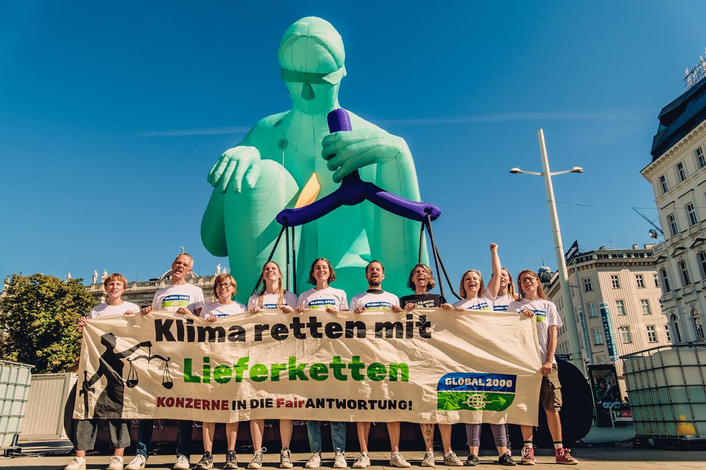 Aktivist:innen halten einen Banner in der Hand mit der Aufschrift "Klimaretten mit Lieferketten". Sie stehen vor einer 10 Meter großen Statue, die Justitia darstellt.