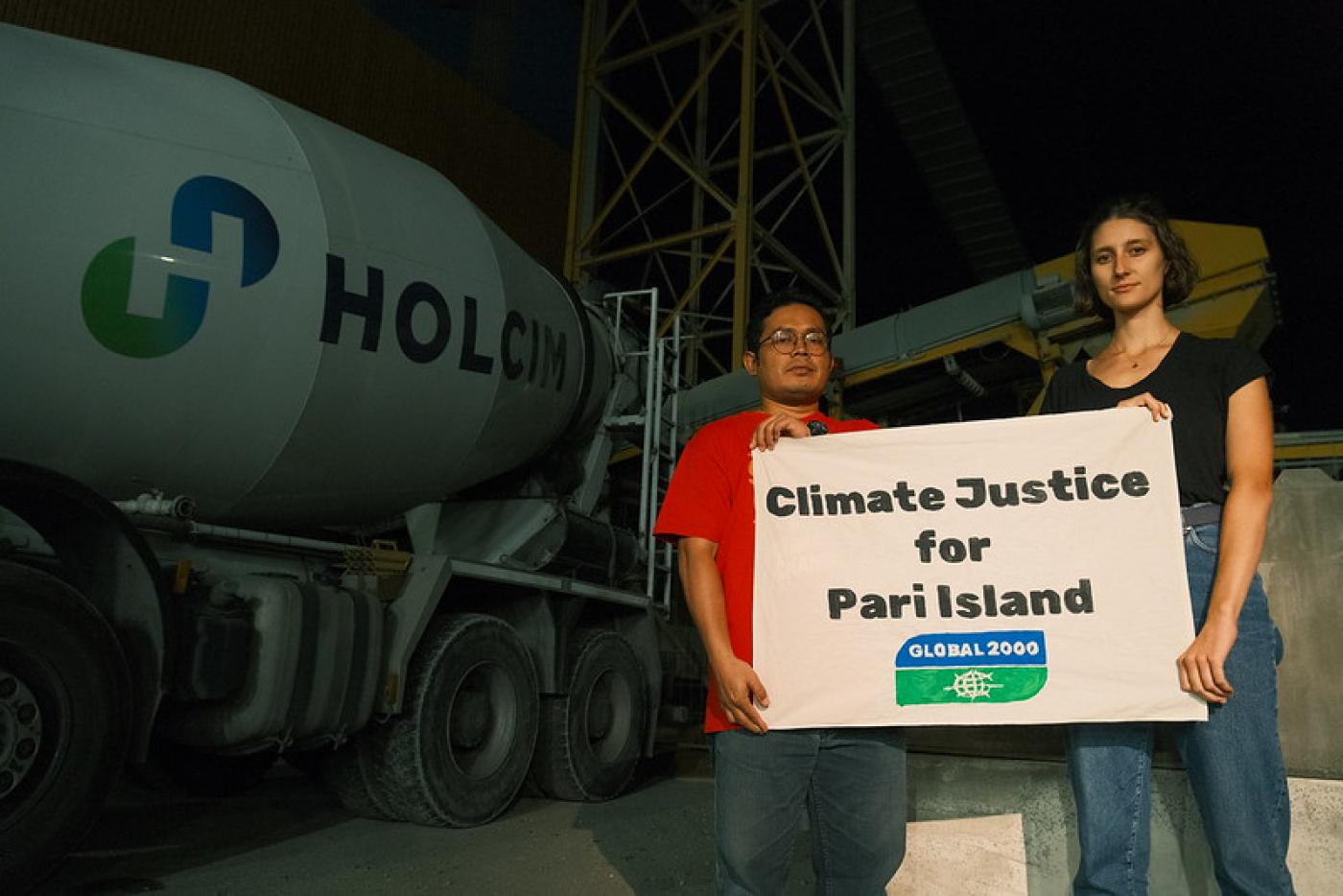 Anna Leitner und Parid stehen vor der Firma Holcim und halten ein Schild in die Kamera, darauf steht "Climate Justice for Pari Island"