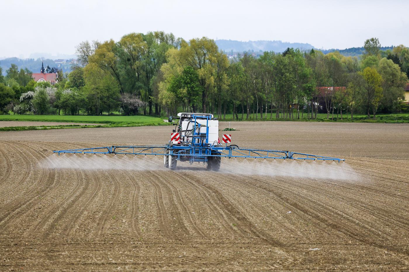 Traktor spritzt Pestizide auf einem Feld. Im Hintergrund sieht mal eine Siedlung. 