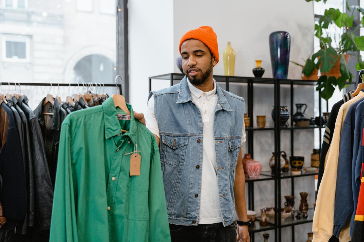 Mann mit oranger Haube und Jeansjacke hält ein grünes Hemd in der Hand und schaut es an. Er steht in einem Vintage store.