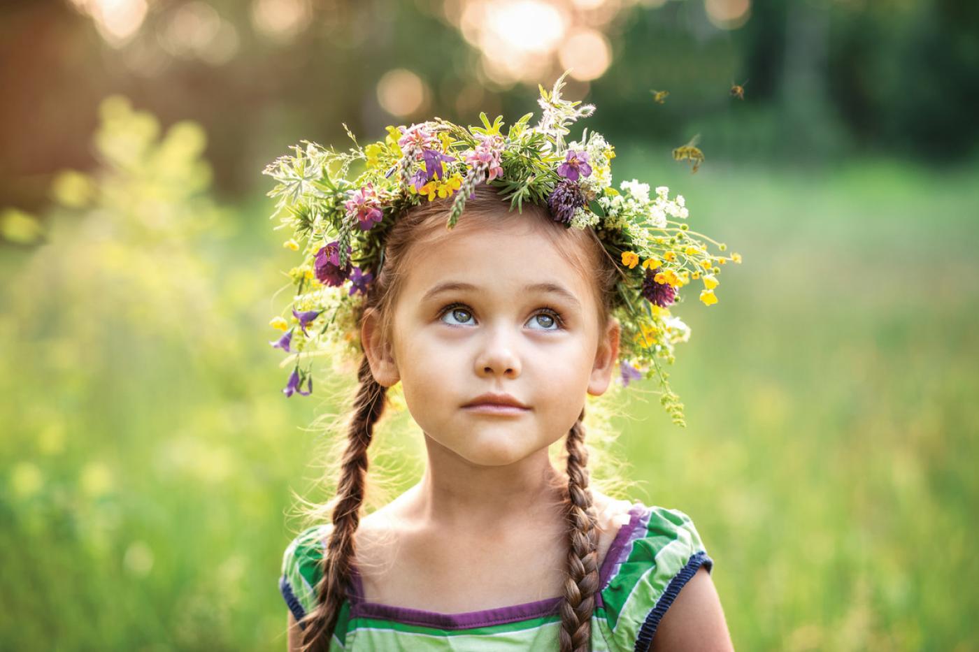Mädchen mit Zöpfen und Blumenkranz schaut rauf in den Himmel. Im Hintergrund sieht man eine weitere grüne Wiese.