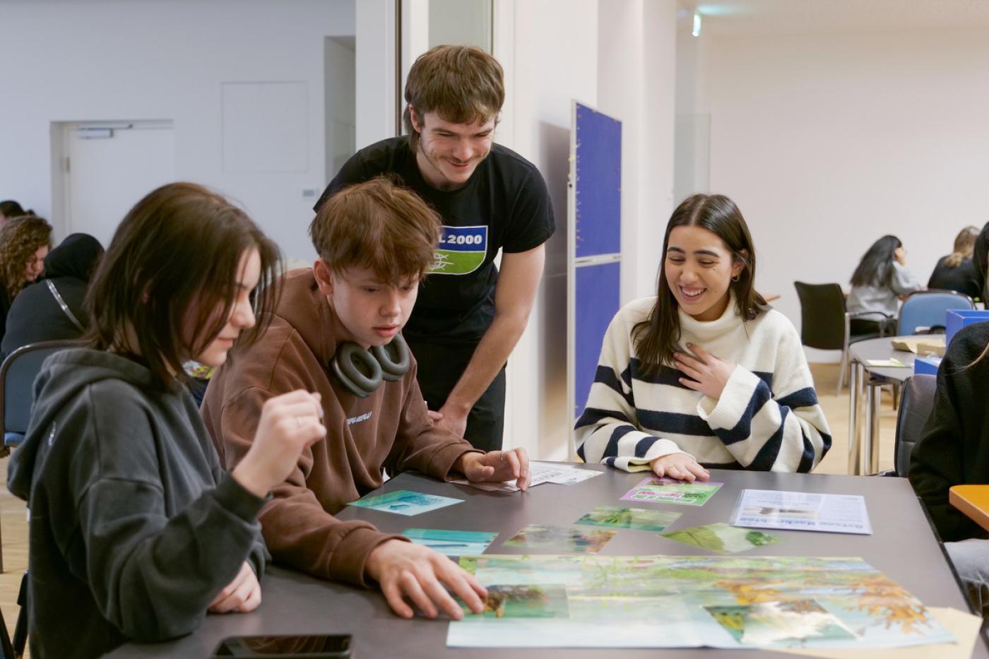 Ein junger Mann mit GLOBAL 2000 T-Shirt beugt sich über einen Tisch, an dem drei Schüler:innen sitzen und an einer Aufgabe arbeiten. Alle lachen.