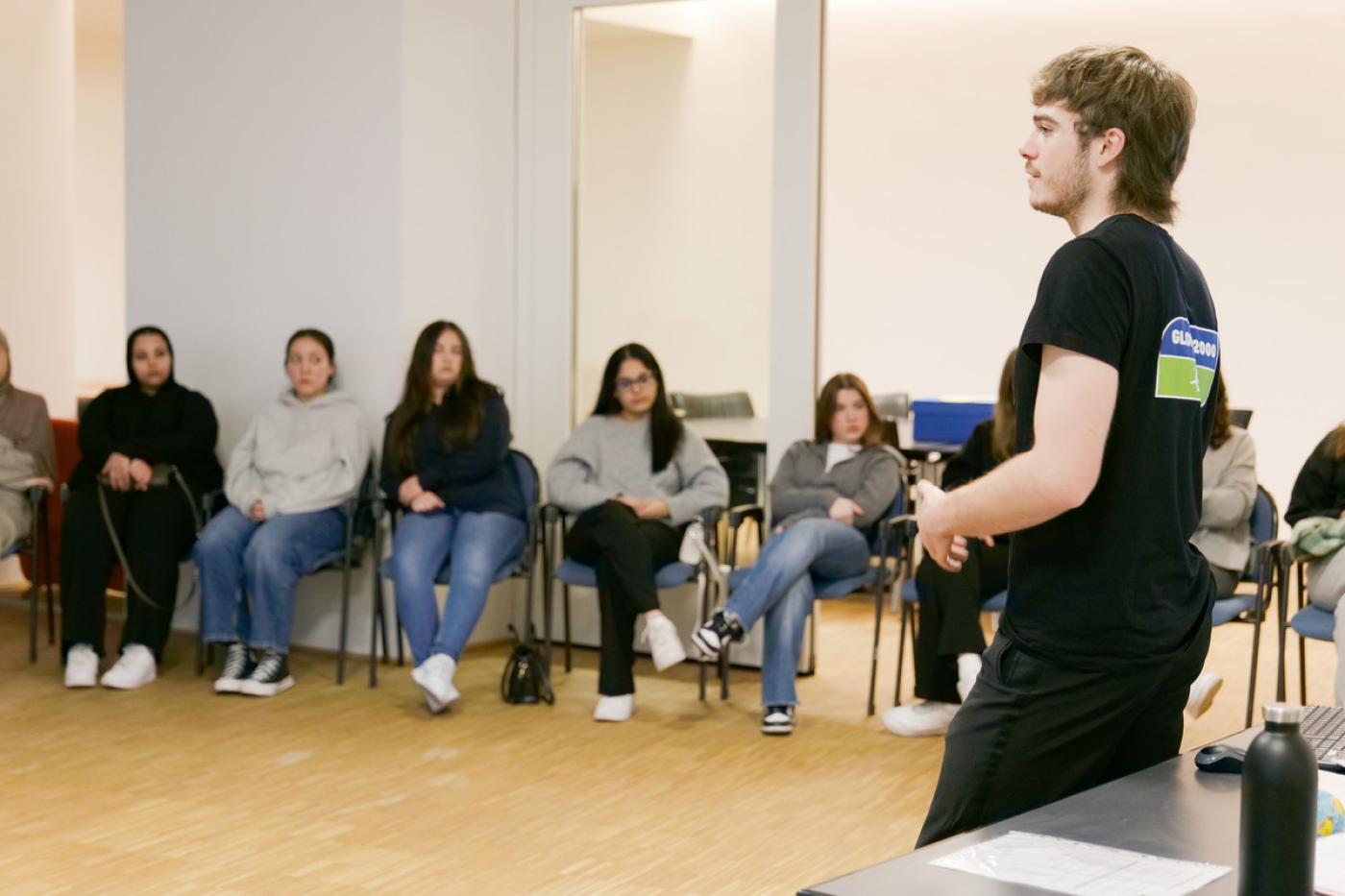 Junger Mann in GLOBAL 2000 T-Shirt steht vor einem Schulklasse im Sesselkreis. Er redet, sie schauen ihn interessiert an. 