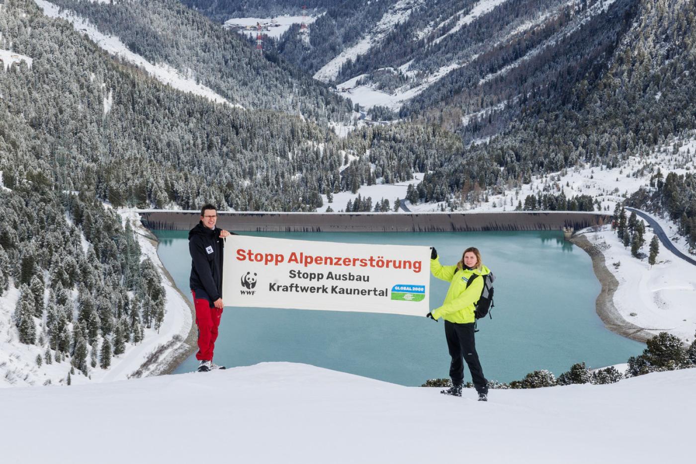 Zwei Aktionist:innen stehen über dem Kraftwerk Kaunertal und halte einen Banner. Darauf sreht: "Stopp Alpenzerstörung"