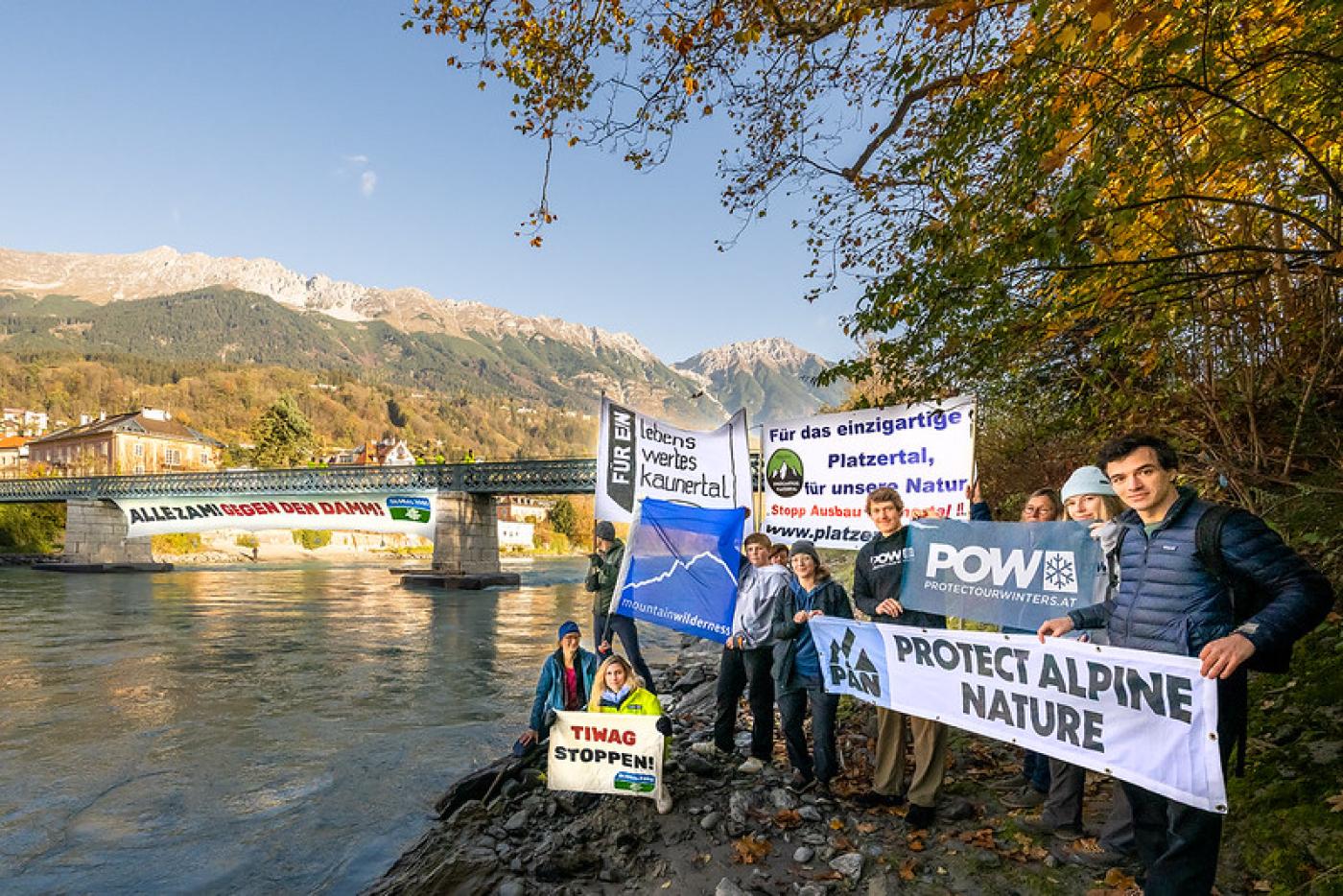 Lokale Innitativen stehen neben dem Inn mit Plakaten, im Hintregrund sieht man das große Banner, dass über der Brücke hängt