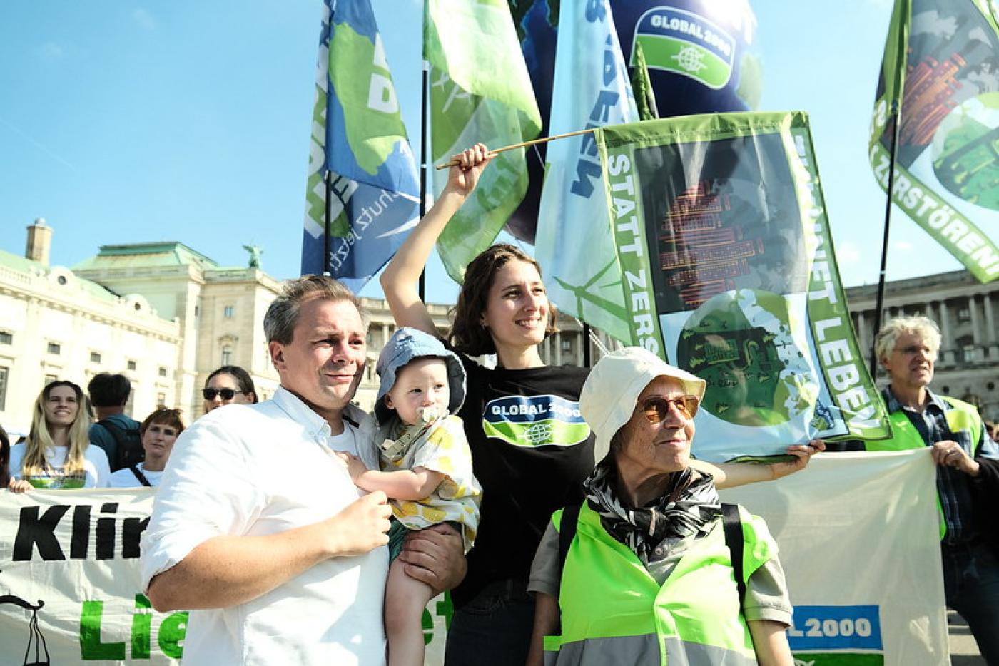 Eine Frau, ein Mann mit Baby im Arm und eine ältere Dame stehen gemeinsam bei einer Demo zusammen und blicken hoffnungsvoll in die Ferne, während die Frau eine Fahne schwenkt.