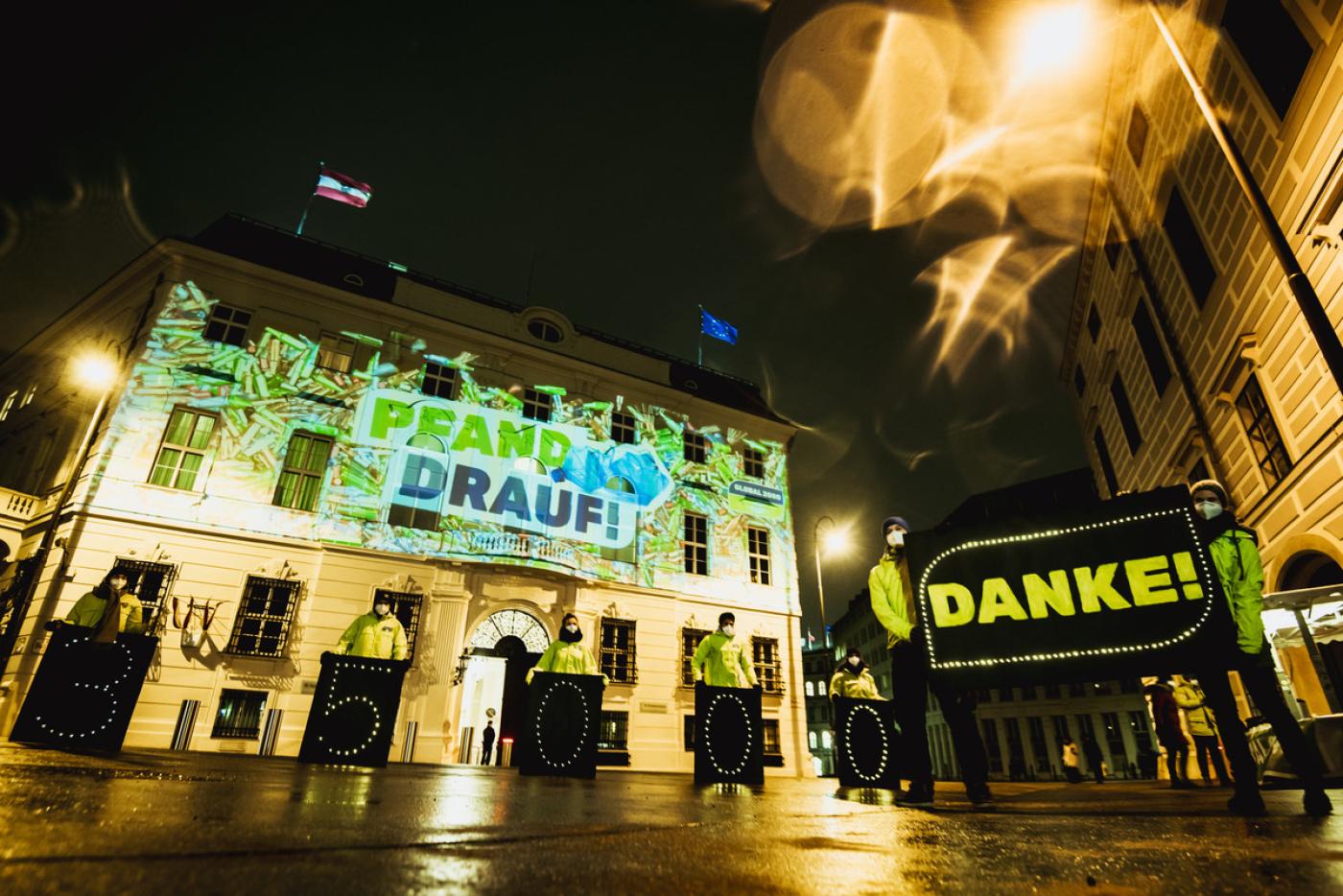 Beamer-Aktion mit dem Pfand-drauf-Logo. Davor stehen Aktivist*innen mit einem Banner, auf dem "Danke" steht.