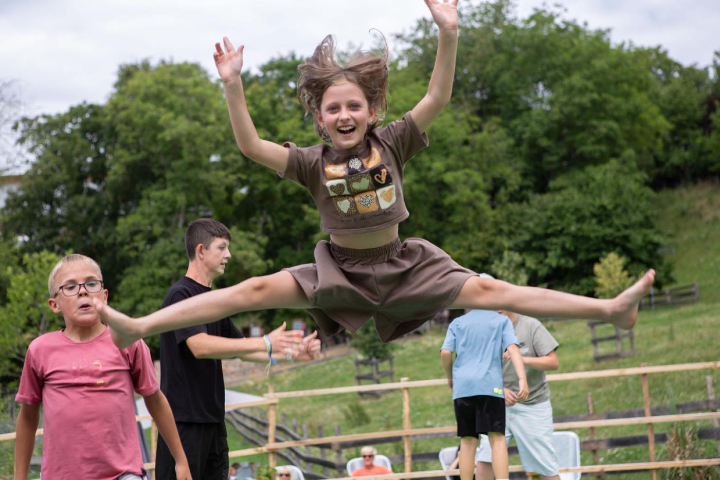 Ein lachendes Mädchen in einem braunen T-Shirt hüpft draußen auf einem Trampolin und macht dabei begeistert einen Spagatsprung in der Luft, mit erhobenen Armen und wehenden Haaren, während andere Kinder im Hintergrund vor einer Wiese mit Bäumen ebenfalls springen und spielen.