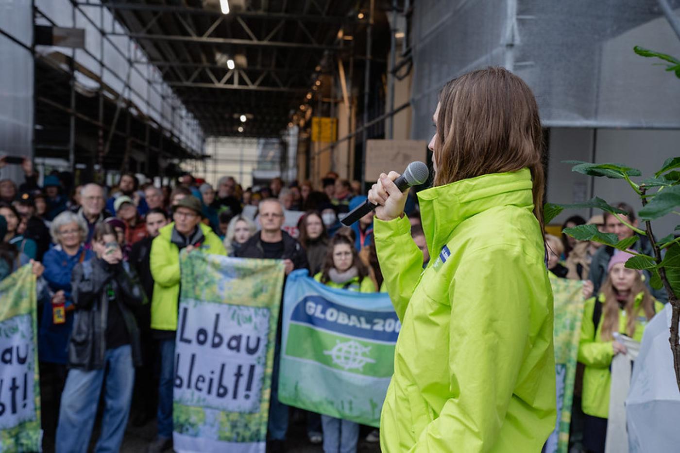 Eine Person in neongelber Jacke, Hannah Keller Klimasprecherin von Global2000, spricht mit einem Mikrofon zu einer Menschenmenge. Die Zuhörer:innen stehen dicht gedrängt unter einem Baugerüst und halten Banner hoch, unter anderem mit den Aufschriften ‚Lobau bleibt!‘ und ‚GLOBAL 2000‘. Die Stimmung ist aufmerksam und engagiert.