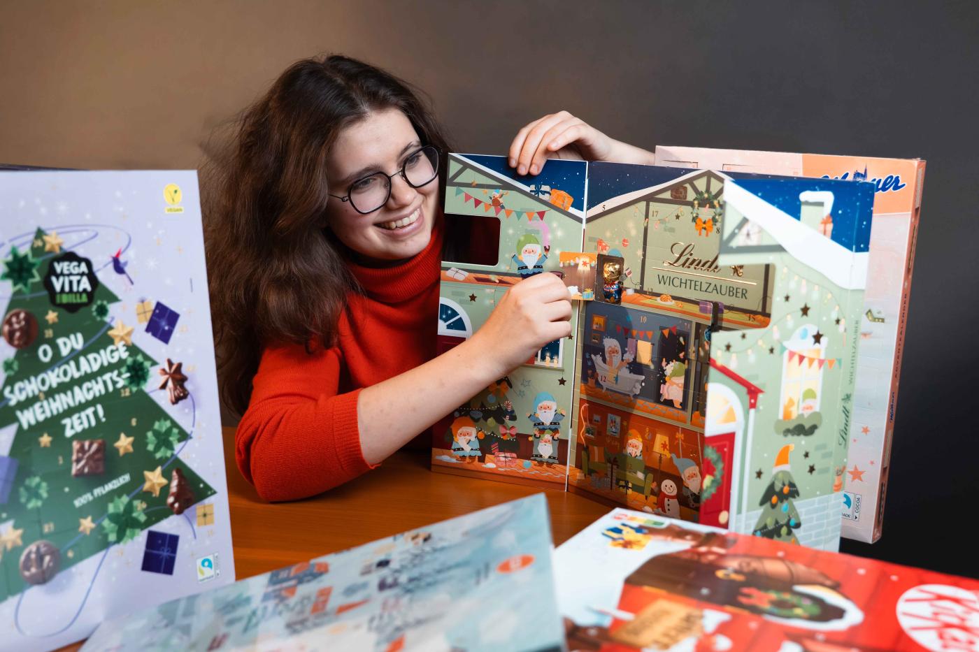 Mädchen in rotem Pulli öffnet Fenster von Schoko-Adventkalender, rundherum mehrere Adventkalender