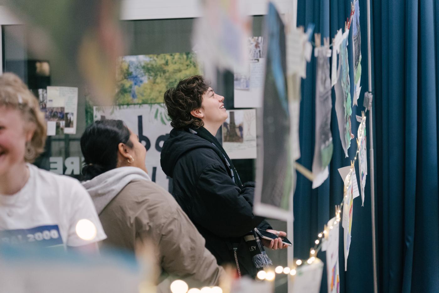 Zwei Personen stehen in einem Innenraum, lächeln und betrachten Fotos und Kunstwerke, die an Schnüren mit Wäscheklammern hängen. Im Hintergrund sind blaue Vorhänge und ein Fenster zu sehen. Lichterketten hängen entlang der Ausstellung.