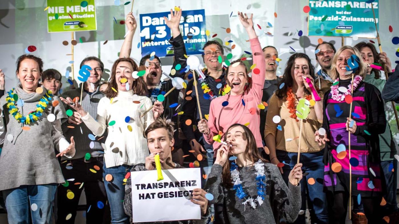 GLOBAL 2000 Mitarbeiter:innen jubeln, es fliegt Konfetti und sie halten Schilder in den Händen wie zB Transparenz hat gesiegt
