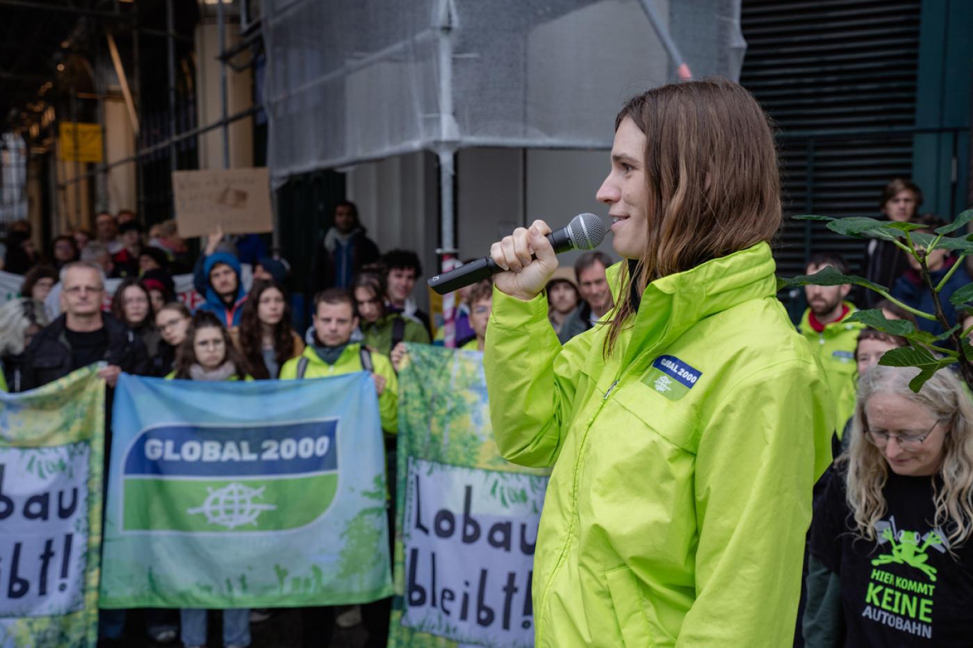 Hannah Keller spricht vor Demonstrant:innen, die Banner halten mit der Aufschrift Lobau bleibt und GLOBAL 2000