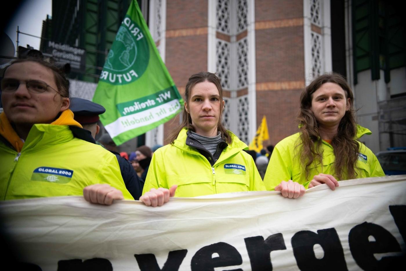 Mehrere Personen in knallgelben Jacken und GLOBAL 2000-Logo mit Banner in den Händen, dahinter Fahnen, auf einer Demo