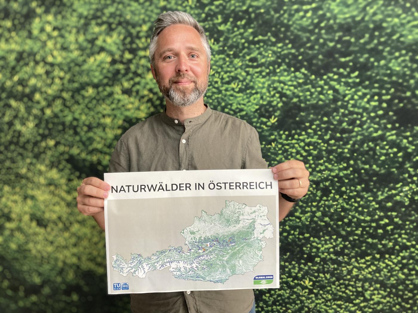 Dominik Linhard, Biodiversitäts-Experte bei GLOBAL 2000, hält einen Ausdruck "Naturwälder in Österreich" mit Österreichkarte und lächelt vor einer Wand mit grünen Blättern