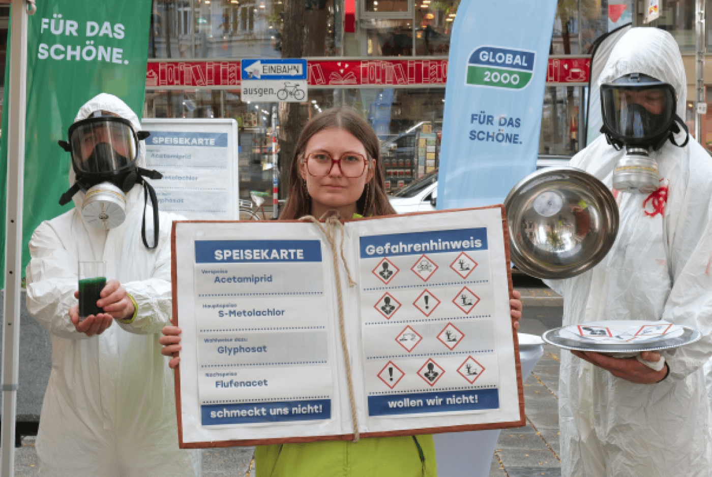 Zwei Personen in Schutzanzügen mit Atemschutzmasken halten links ein grünes Getränk und rechts eine Metallschüssel, dazwischen eine Frau mit Brille hält ein großes Schild mit einer Speisekarte und Gefahrensymbolen, im Hintergrund ein Banner mit der Aufschrift 'GLOBAL 2000 FÜR DAS SCHÖNE'