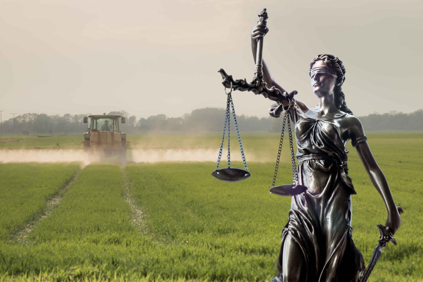 Statue der Justitia mit Waage im Vordergrund vor einem Traktor, der auf einem Feld Pestizide versprüht