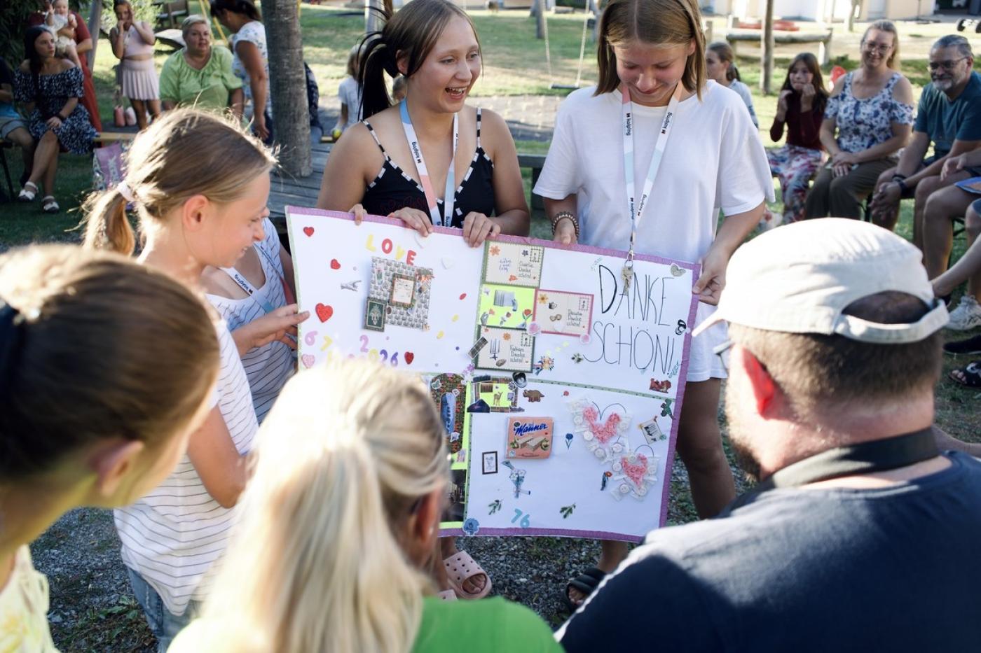 Drei junge Frauen zeigen im Freien ein großes, bunt gestaltetes Plakat mit der Aufschrift 'DANKE SCHÖN' vor einer Gruppe sitzender und stehender Menschen.