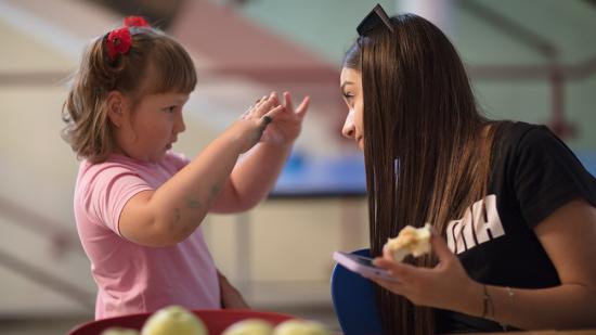 Ukrainisches Mädchen formt Herz mit ihren Fingern und zeigt es der Projekt-Mitarbeiterin Valeriia, während diese einen Apfel isst