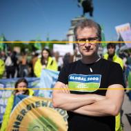 Johannes Wahmüller, Klima- & Energie-Sprecher bei GLOBAL 2000 steht mit verschränkten Armen und entschlossenem Blick vor für Umwelt- und Klimaschutz protestierenden Menschen