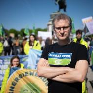 Johannes Wahlmüller steht mit schwarzem T-Shirt mit Aufdruck "GLOBAL 2000" vor einer Gruppe von Menschen mit bunten Bannern bei einer Demonstration im Freien