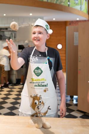 Junge mit Schürze und Kopfbedeckung schupft lachend Teig beim backen.