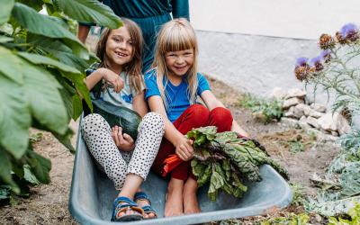 Zwei Mädchen sitzen in einer Scheibtruhe und halten Zucchini und Mangold in der Hand