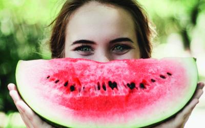 Eine braunhaarige Frau versteckt ihr Gesicht halb hinter einem Wassermelonenstück, das sie vor ihr gesicht hält.