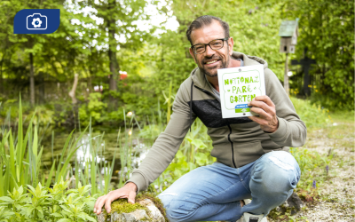 Karl Ploberger hält Nationalpark Garten Plakette in der Hand und kniet in seinem Garten