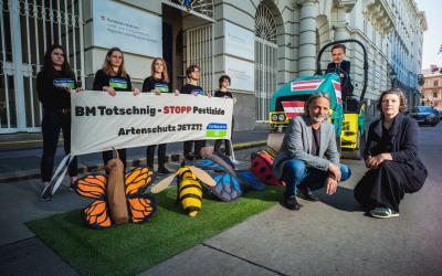 Totschnig fährt mit einer Walze über Bestäuber. Im Hintergrund stehen Aktivist:innen mit einem Plakat.