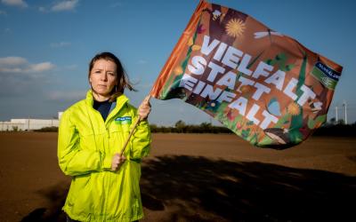 Brigitte Reisenberger steht auf einen braunen Feld und hält eine Fahne in der Hand auf der steht "Vielfalt statt Einfalt"