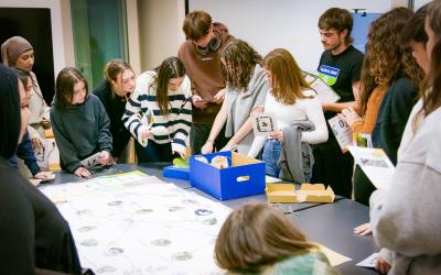 Schülergruppe steht um einen Tisch und sie schauen interessiert in eine blaue Box. Einige von ihnen halten eine Karte in den Händen. 
